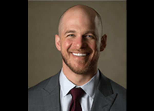 Smiling man in a gray suit and burgundy tie, facing the camera, with a neutral background
