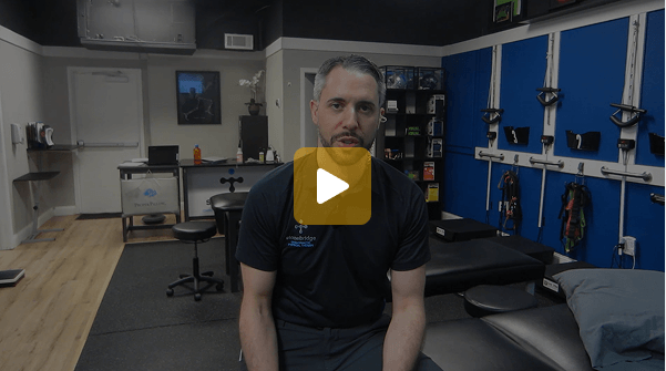 Video thumbnail of a man seated in a therapy clinic with exercise equipment on blue wall panels behind him, play button overlayed