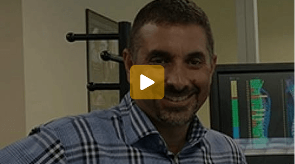 Video thumbnail: smiling man in a blue plaid shirt in an office, with a computer monitor in the background; play button centered.