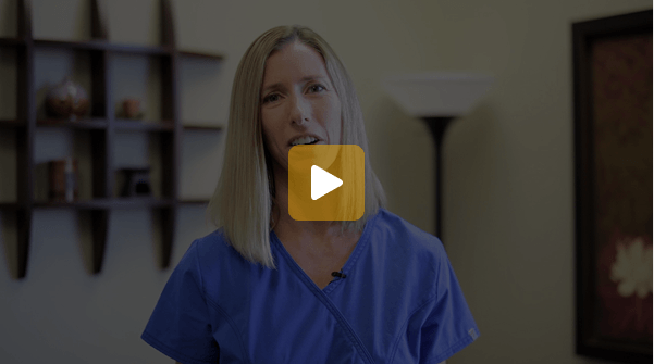Video thumbnail: woman in blue scrubs speaking to the camera in a room with shelves and a lamp behind her.