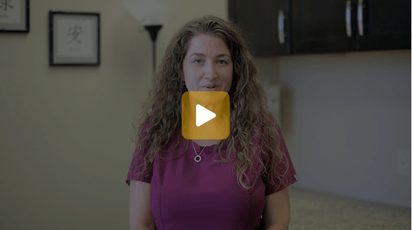Video thumbnail: woman with long curly hair in a magenta shirt speaks to the camera in a kitchen; play button overlay appears in the center.